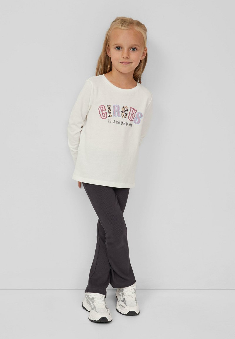 Junges Mädchen mit blonden Haaren, das ein weißes Langarmshirt mit dem Text "Der Zirkus ist um mich herum", dunkle Hose und weiße Sneakers trägt, steht vor einer grauen Wand.