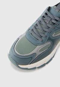 Tênis com design em camadas em tons de azul e verde, apresentando uma combinação de materiais lisos e texturizados, e sola de borracha branca.