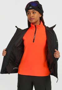 Zwarte jas geopend om een feloranje pullover te onthullen, met een kwart rits. De pullover is gemaakt van fleece materiaal.