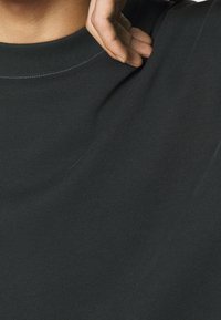 Sweat-shirt gris foncé à col rond avec un tissu texturé. Un gros plan montre le col et les détails de couture, avec une main tenant le tissu.