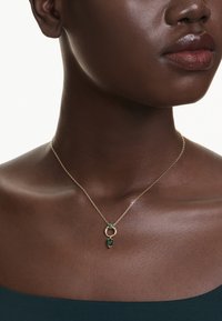 Close-up van een persoon die een gouden ketting draagt met een cirkelvormige hanger en twee groene edelstenen op een donkerblauwe top.