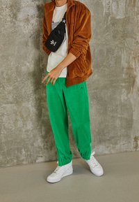 Person som bär en brun jacka, gröna byxor, vita sneakers och en svart Adidas crossbody-väska, stående mot en betongvägg.