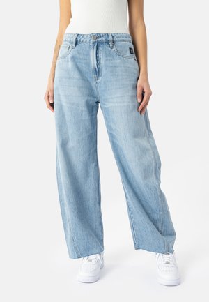 Person iført lyseblå vidde jeans, hvide sneakers og en hvid ærmeløs top, stående foran en ensfarvet baggrund.