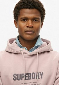 Lichtroze hoodie met een trekkoord waarin het "SUPERDRY" logo in donkergrijs is gedrukt. Onderaan is een denimhemdkraag zichtbaar. Soepele stoftextuur.
