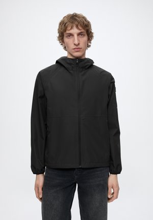 Junger Mann mit lockigem Haar, der eine schwarze Kapuzenjacke und dunkle Jeans vor weißem Hintergrund trägt.