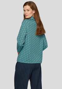 Türkis gemustertes Hemd mit geometrischen Designs und hohem Kragen, kombiniert mit dunkler, gestreifter Hose. Glatte Stoffstruktur.