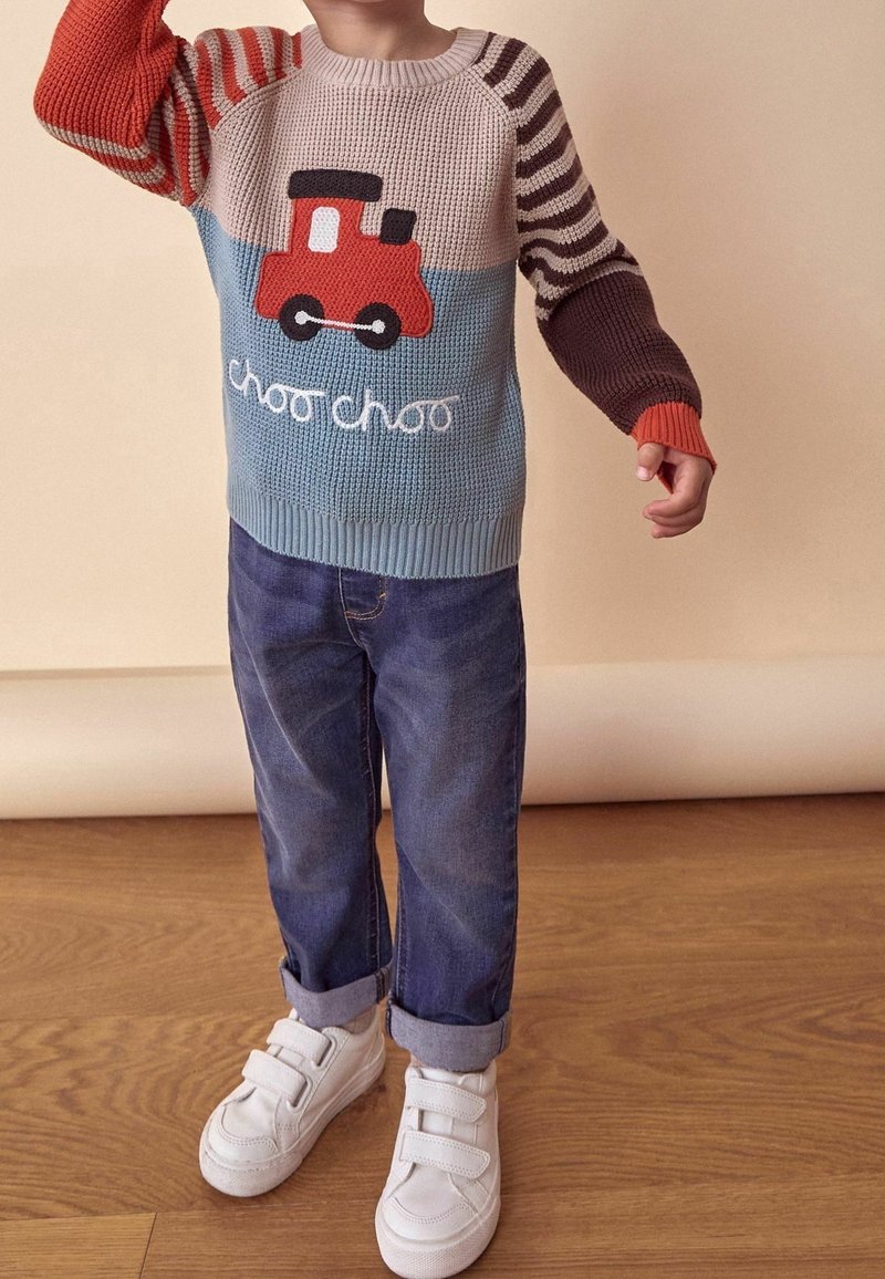 Enfant portant un pull coloré tricoté avec un train rouge et le texte « choo choo », un jean bleu retroussé aux chevilles, et des baskets blanches à scratch.