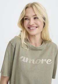 Camiseta verde oliva hecha de una tela suave, con la palabra "amore" en letras brillantes y brillantes en el pecho. Mangas cortas, corte relajado.