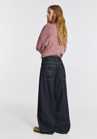 Túlméretezett rózsaszín kötött pulóver és sötétkék, széles szárú farmer, melynek hátsó zsebein szív hímzés található. A modell háttal áll a kamerának.