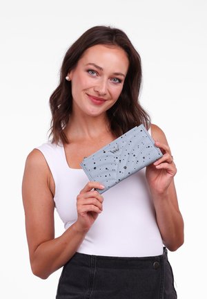 Light blue wallet with quilted design and black speckled accents, featuring a silver logo and zip closure, held in a hand.