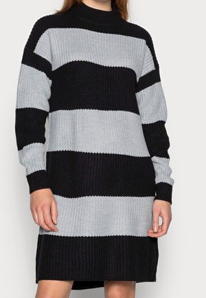 Personne portant une robe pull en tricot rayée noire et grise, longue, avec des manches longues et un col montant.