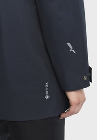 GORE-TEX-jacka i djup marinblå, med en slät textur, en silverfärgad logotyp på ärmen och tryckknappsstängning vid manschetten.