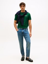 Zeleni bombažni majica s logotipom, mornarsko modri pulover okoli ramen, modri jeans in črne loafer čevlje, ki stoji ob beli ozadju.