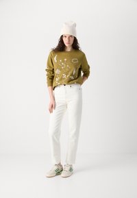 Sweatshirt vert olive avec broderies florales blanches, associé à un jean droit blanc et des baskets beiges avec des accents verts, surmonté d'un bonnet crème.