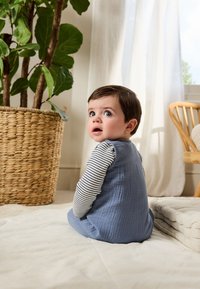 Vêtement sans manches bleu clair texturé porté sur une chemise à manches longues blanche et rayée marine, enfant assis sur un tissu clair dans une pièce lumineuse.