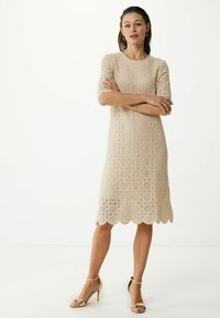 Beige spetsklänning med korta ärmar, vågig fåll och blommönster. Matchas med gyllene högklackade sandaler.