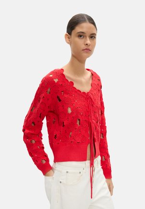 Kobieta w czerwonym, szydełkowanym kardiganie z kwiatowym wzorem i wiązaniem z przodu, w białych spodniach, stojąca na jednolitym tle.