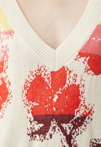 Pull en tricot avec un col en V, présentant un motif floral en rouge, orange et rose sur un fond crème. Texture côtelée autour de l'encolure.