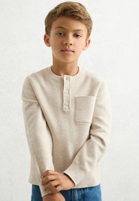 Beige ribgebreide blouse met lange mouwen en een henley-halslijn, voorzien van drie knopen en een borstzak, gecombineerd met blauwe spijkerbroeken.