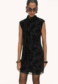 Woman wearing sleeveless black floral velvet dress with neck tie, dark sunglasses, gold hoop earrings, bangles, and black sheer tights.