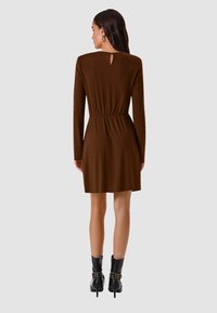 Vestito marrone a maniche lunghe con vita aderente e dettaglio cut-out sulla schiena, abbinato a stivaletti neri con borchie. Tessuto liscio e lunghezza al ginocchio.