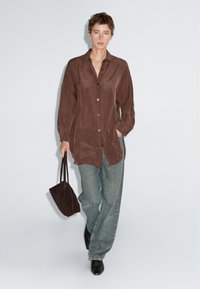 Brun ruskindsskjorte med knapper, lange ærmer, store forlommer og en løs pasform, kombineret med lyseblå vidde-ben jeans og en lille sort taske.