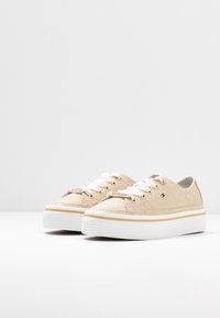 Sneakers in tela beige con lacci bianchi, occhielli dorati e superficie texturizzata. Suola in gomma bianca con striscia dorata attorno alla base.