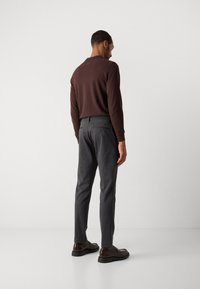 Hombre vestido con una camisa de manga larga marrón, pantalones slim-fit de color gris oscuro y mocasines de cuero marrón, de pie frente a una pared blanca.