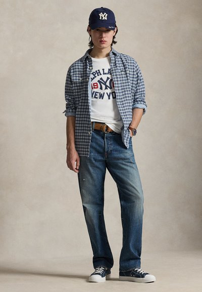 Mannequin portant une chemise à carreaux bleue sur un t-shirt blanc à motif graphique, un jean bleu et des baskets bleu marine, coiffé d'une casquette bleu marine avec un logo.