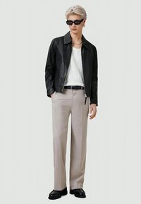 Pessoa a usar uma jaqueta de couro preta, uma camisa branca, umas calças bege, um cinto preto, sapatos pretos e óculos de sol escuros, de pé contra um fundo liso.