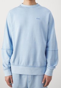 Man med ljusblå sweatshirt och matchande byxor, med "BOSS"-logotyp broderad på övre bröstet.