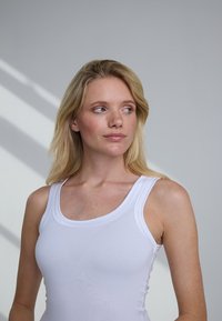 Blonde vrouw in een wit hemd kijkt zijwaarts met een neutrale uitdrukking tegen een effen grijze achtergrond met licht en schaduw.