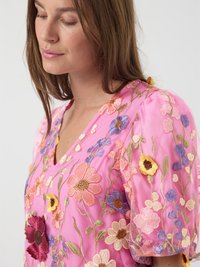Rosa blus med blomsterbroderi och korta puffärmar. Har färgglada blommor i olika nyanser och intrikat broderi på ett genomskinligt tyg.