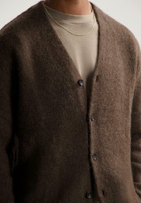 Bruine, fuzzy cardigan met een V-hals ontwerp, voorzien van drie donkere knopen en zijzakken, gelaagd over een lichtgrijs shirt.