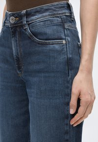 Κοντινό πλάνο ενός ατόμου που φοράει σκούρο μπλε τζην με πορτοκαλί ραφές και καφέ μπλούζα, το χέρι ακουμπάει δίπλα στον μηρό.