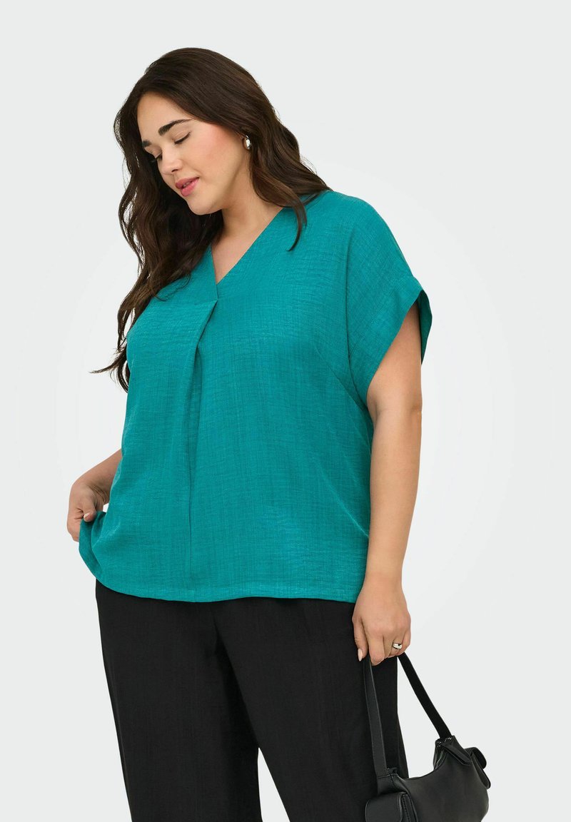 Femme aux longs cheveux foncés portant un haut turquoise à manches courtes et un pantalon noir, tenant un sac à main noir d'une main.