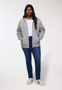 Felpa grigia con zip e coulisse, con due tasche frontali, indossata sopra una t-shirt bianca. Abbinata a jeans blu scuro e sneakers beige.