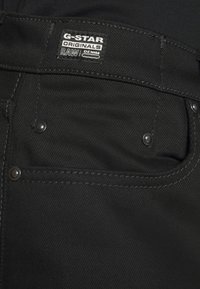 Jeans in denim nero con etichetta G-Star, caratterizzati da rivetti metallici, cuciture a contrasto e una consistenza liscia. Tasche visibili nella parte anteriore.