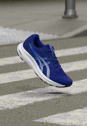 Scarpa da corsa blu con suola bianca e logo, a mezz'aria sopra un attraversamento pedonale, che mostra movimento e ammortizzazione sull'asfalto.