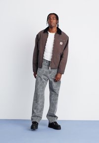Brun zip-jakke med svart krage, hvit t-skjorte og grå camo-mønstrede jeans. Båret med svarte sneakers, stående på blå bakgrunn.