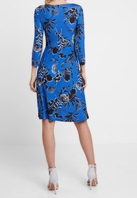 Donna che indossa un vestito blu a ginocchio con motivi floreali in bianco e nero e scarpe con tacco alto bianche a forma di caviglia, in piedi con la schiena rivolta.