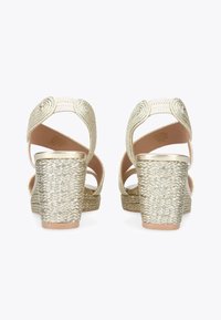 Gold wedge sandals with a woven texture, featuring a rounded strap design and a thick platform heel. The back view displays the strap detailing.