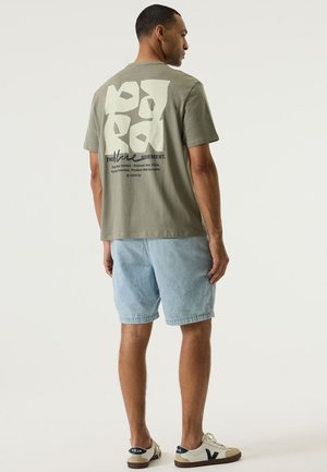 Olivegrünes Baumwoll-T-Shirt mit weißem abstraktem Design auf der Rückseite, kombiniert mit hellblauen Denim-Shorts. Das Modell trägt beigefarbene und weiße Turnschuhe.