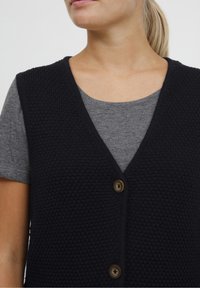 Granatowa dzianinowa kamizelka o dekolcie w kształcie litery V, z fakturowanej tkaniny i drewnianymi guzikami, noszona na szarej koszulce.