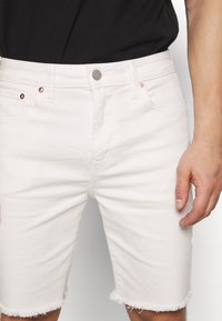Bílé denimové šortky s třepeným lemem, dvěma předními kapsami a měděným kováním. Hladká textura a přiléhavý střih.