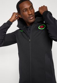 Férfi, aki éppen a kapucnis fekete cipzáras dzseki kapucniját igazítja, amelyen zöld és piros sportlogó látható a mellrészen, egyszerű fehér háttér előtt.