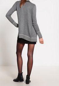 Schwarz-weiß gestreifter langer Cardigan mit Schalkragen, figurbetontem Schnitt und kontrastierendem Saum, kombiniert mit einem schwarzen Rock und Stiefeletten.