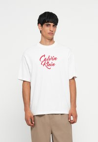 Camiseta de algodón blanca de corte holgado, con un logotipo rojo en script "Calvin Klein" en el frontal. Diseño de mangas cortas y cuello redondo.