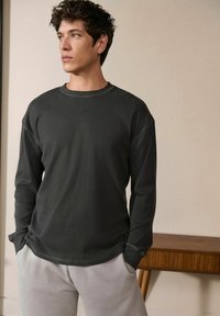 Donkergrijs langemouwen T-shirt met een geribbelde textuur, ronde halslijn en verlaagde schouders; gedragen met lichtgrijze joggingbroek.