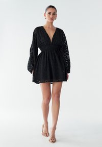 Juoda nėriniuota suknelė su ilgomis pūstomis rankovėmis, giliais V formos iškirptėmis ir elastingu juosmeniu. Sijonas sluoksniuotas, pusiau permatomas.